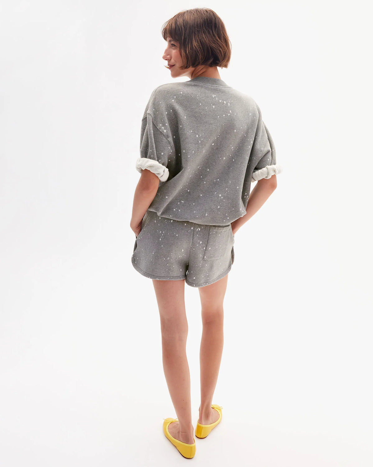 A person with short brown hair and yellow flats stands against a white background, wearing the Clare Vivier SS Oversized Sweatshirt— a loose-fitting, gray speckled 100% cotton top with rolled sleeves, paired with matching shorts.