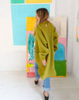 Kerri Rosenthal Sienna Duster in olive alpaca blend worn with blue jeans against colorful abstract art.