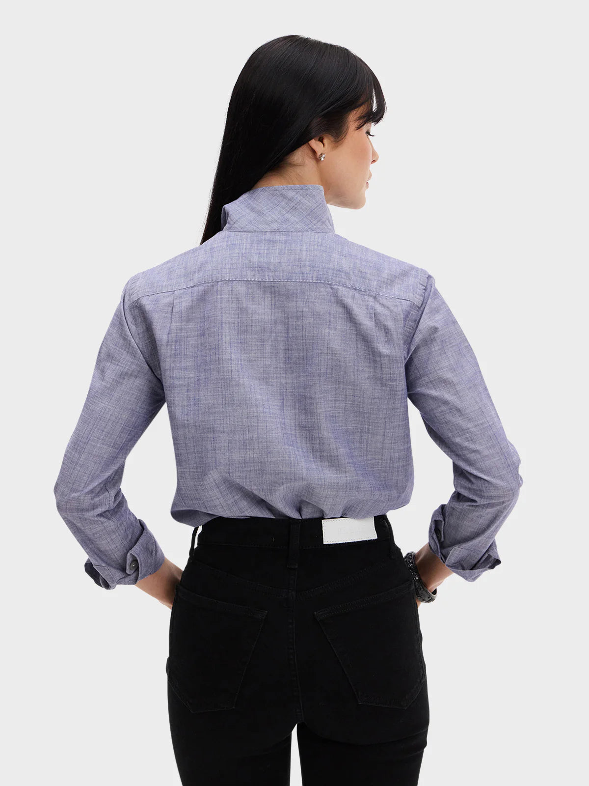 A woman with long dark hair is seen from behind, hands on hips, wearing the Denim Country Club shirt by Sarah Alexandra, LLC—light purple chambray with a one-piece collar and rolled-up sleeves—paired with black jeans against a plain background.