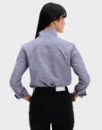 A woman with long dark hair is seen from behind, hands on hips, wearing the Denim Country Club shirt by Sarah Alexandra, LLC—light purple chambray with a one-piece collar and rolled-up sleeves—paired with black jeans against a plain background.