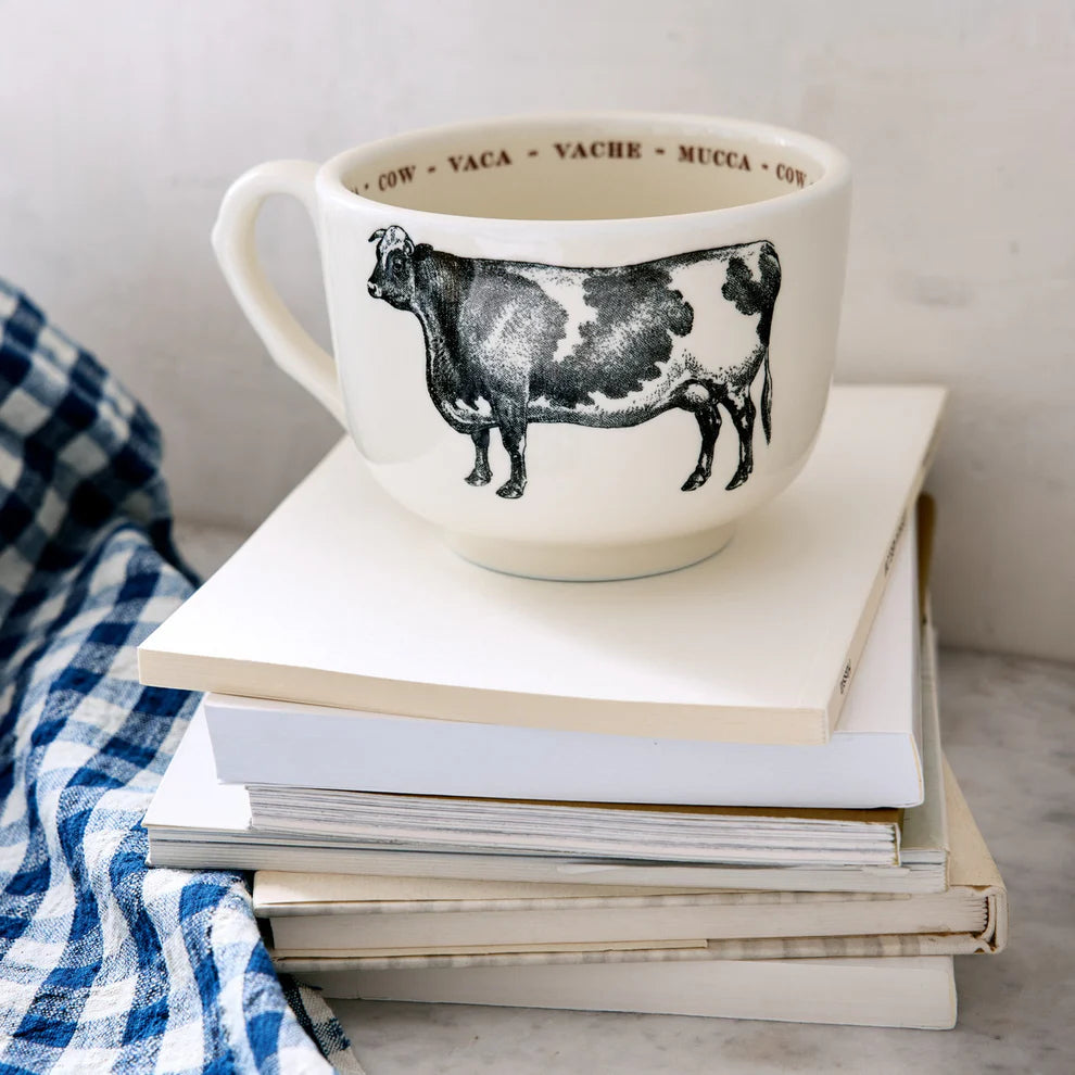 The Grand Cup by Sir/Madam, a large white cup featuring a black cow illustration and "cow" in multiple languages on the rim, sits atop books beside a blue-and-white checkered cloth.