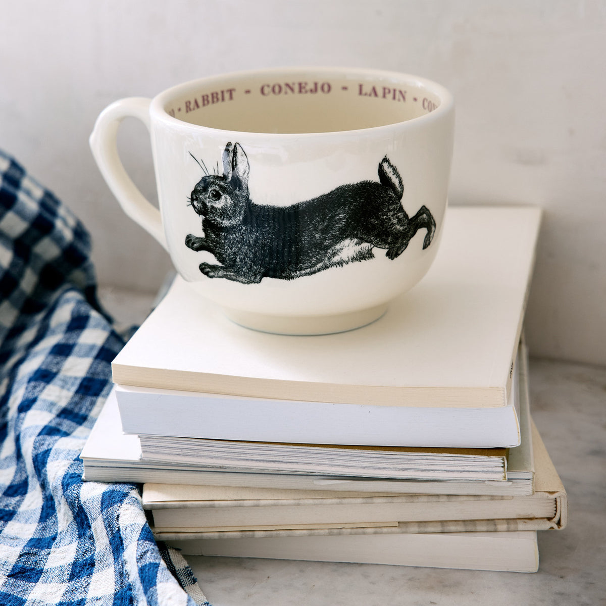 The Grand Cup by Sir/Madam, featuring a black rabbit illustration and "rabbit" in several languages around the rim, sits atop white books next to a blue and white checkered cloth.