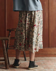 A person wearing a light blue jacket and The Canary Skirt by The Great Inc.—a cream-colored floral silk twill skirt with a smocked waistband—stands indoors near two wooden chairs, with a wooden wall and tiled floor in the background.