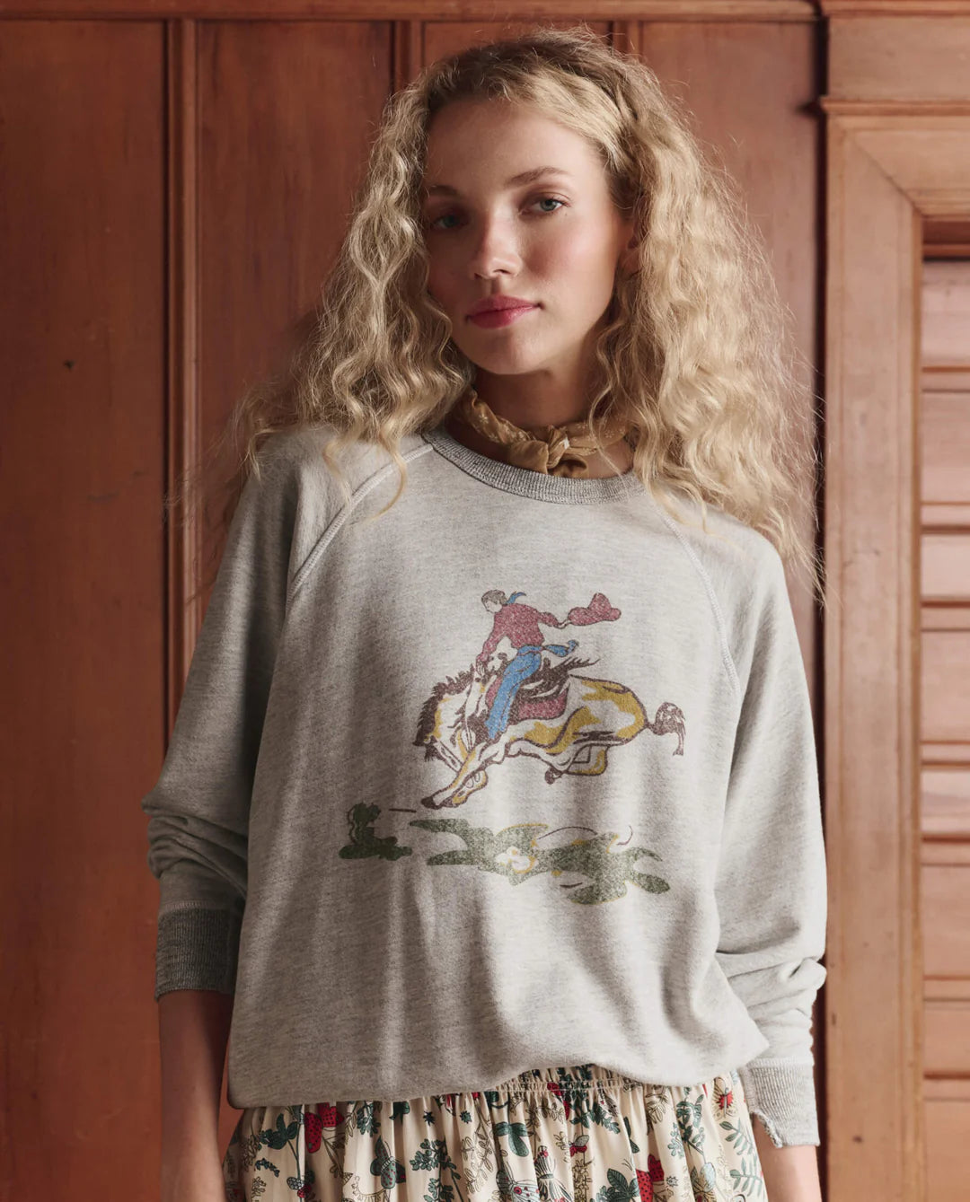 A woman with long, curly blonde hair wears The College Sweatshirt 2026 by The Great Inc.—an oversized vintage washed terry style with a cowboy horse graphic—paired with a floral skirt, standing indoors against wooden paneling.