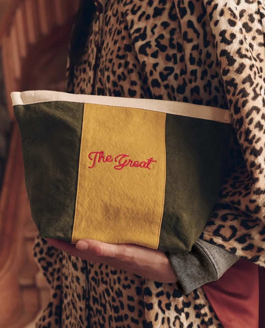 The Striped Pouch in olive green and yellow cotton canvas with a cream band and red logo held in hands.