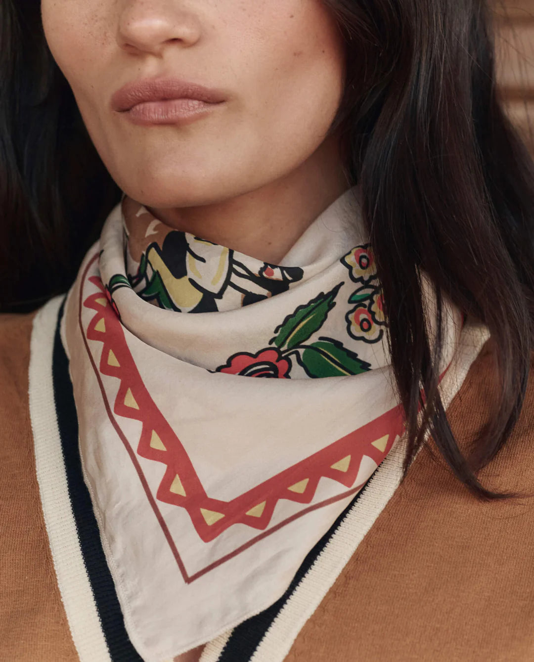 A woman wears The Derby Silk Bandana by The Great Inc., featuring a vintage light floral design around her neck, paired with a tan sweater with black and white trim. Only the lower half of her face and neck are visible.
