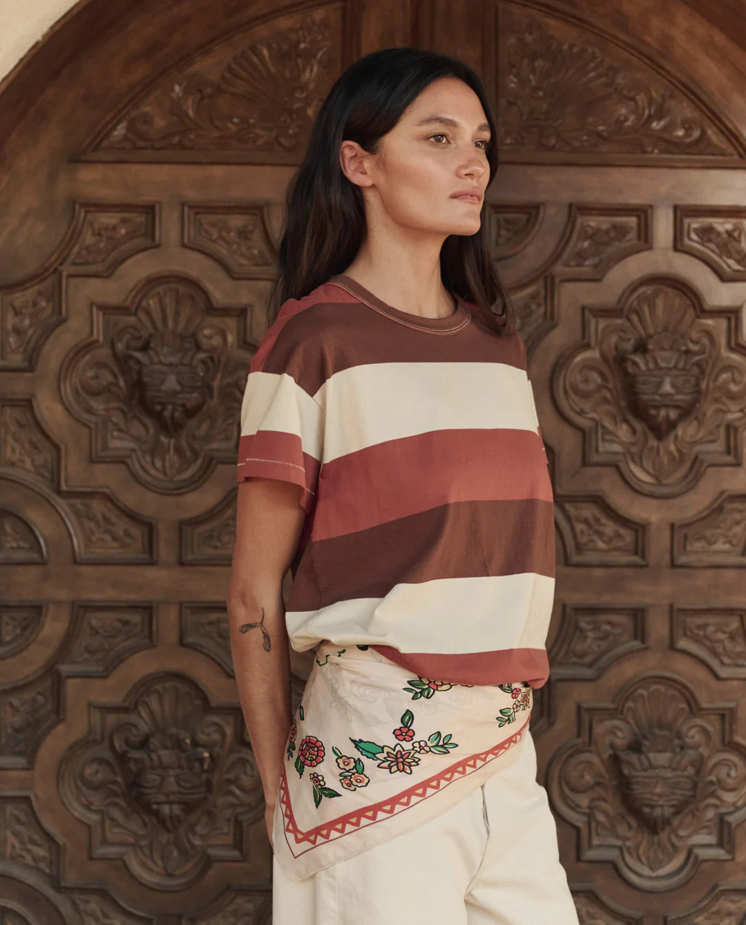 A woman with long dark hair stands before an ornate wooden door, wearing a striped t-shirt, cream floral-embroidered shorts, and The Derby Silk Bandana by The Great Inc. tied at her neck for an American Western touch.