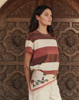 A woman with long dark hair stands before an ornate wooden door, wearing a striped t-shirt, cream floral-embroidered shorts, and The Derby Silk Bandana by The Great Inc. tied at her neck for an American Western touch.