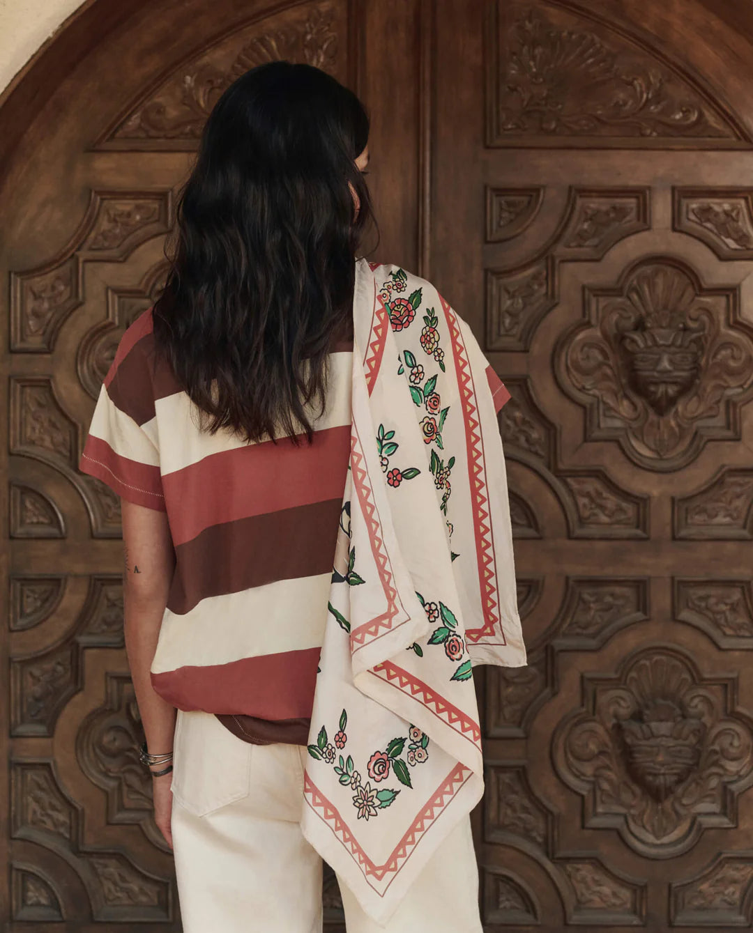 A person with long dark hair stands before ornate wooden doors, wearing a striped shirt, light pants, and The Great Inc.'s The Derby Silk Bandana draped over their shoulder for an American Western-inspired look.