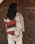 A person with long dark hair stands before ornate wooden doors, wearing a striped shirt, light pants, and The Great Inc.'s The Derby Silk Bandana draped over their shoulder for an American Western-inspired look.