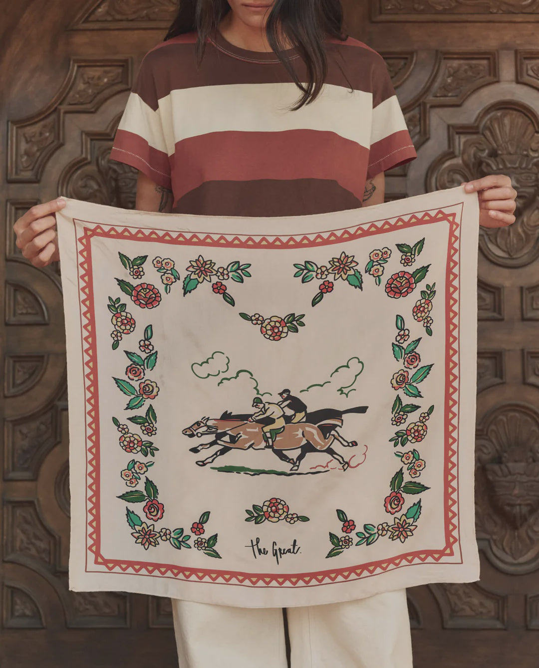 A person in a striped shirt holds The Derby Silk Bandana by The Great Inc.—a square silk scarf with floral borders, jockeys on horses, and "The Great" inscribed at the bottom in classic American Western style.