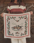 A person in a striped shirt holds The Derby Silk Bandana by The Great Inc.—a square silk scarf with floral borders, jockeys on horses, and "The Great" inscribed at the bottom in classic American Western style.