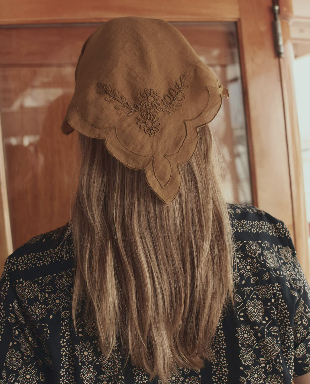 A person with long, light brown hair stands with their back to the camera, wearing The Great Inc.'s Scalloped Bandana and a dark patterned garment in front of a wooden door.