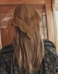A person with long, light brown hair stands with their back to the camera, wearing The Great Inc.'s Scalloped Bandana and a dark patterned garment in front of a wooden door.