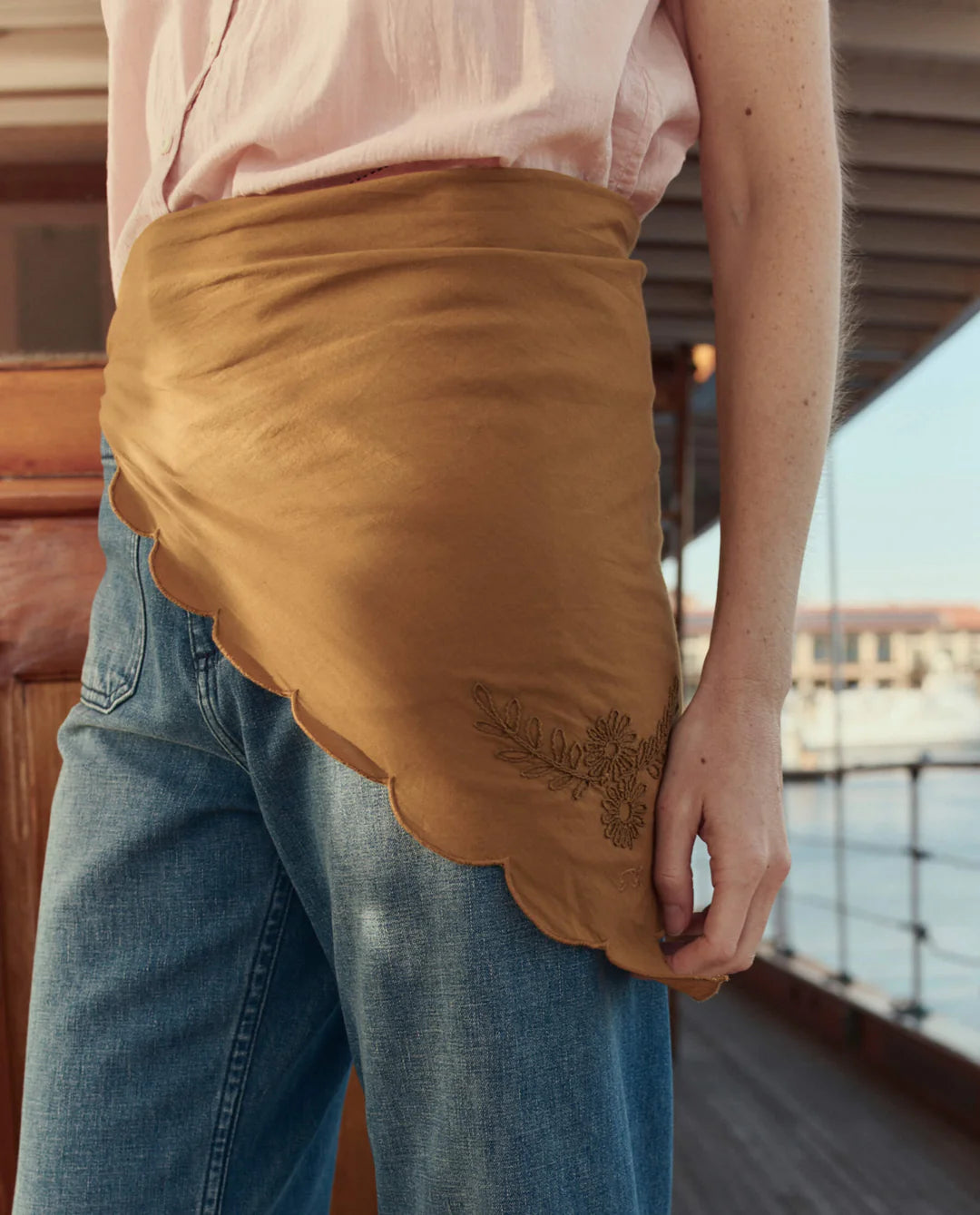 A person stands on a wooden deck near the water, wearing blue jeans, a light pink shirt, and The Scalloped Bandana skirt with vintage handkerchief-inspired floral embroidery by The Great Inc.