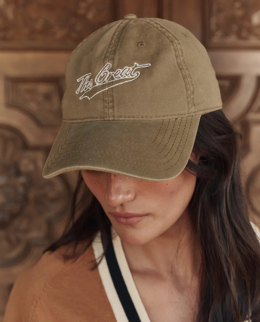 A person wearing The Great Inc. "The Baseball Hat" in khaki with vintage embroidery on the front looks downward, with an ornate wooden background partially visible.