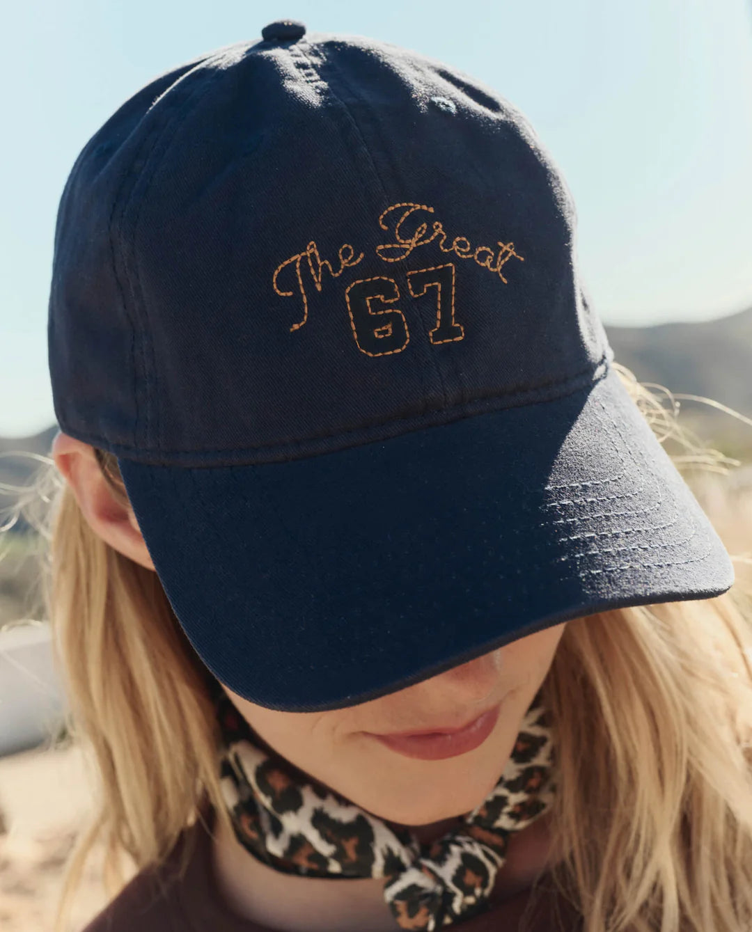 A person wears The Great Inc.'s The Baseball Hat in navy blue with yellow vintage "The Great 67" embroidery and adjustable strap, paired with a leopard print scarf, outdoors in the sun.