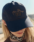A person wears The Great Inc.'s The Baseball Hat in navy blue with yellow vintage "The Great 67" embroidery and adjustable strap, paired with a leopard print scarf, outdoors in the sun.