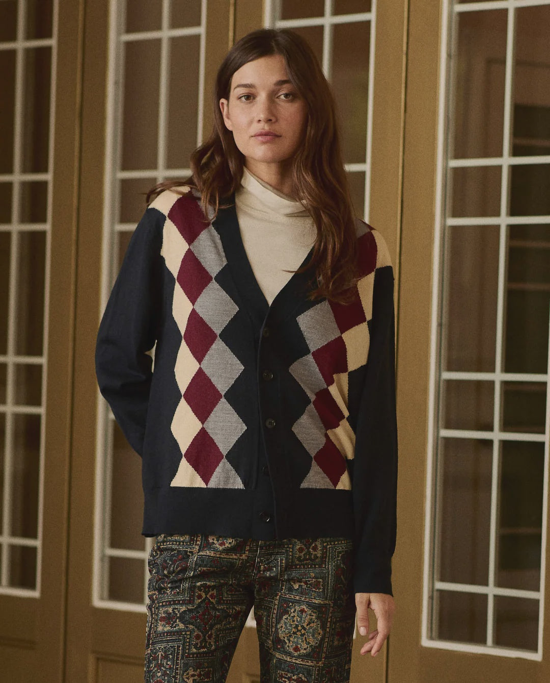 The Argyle Menswear Cardigan in navy, burgundy, and cream worn over a beige mock neck top.