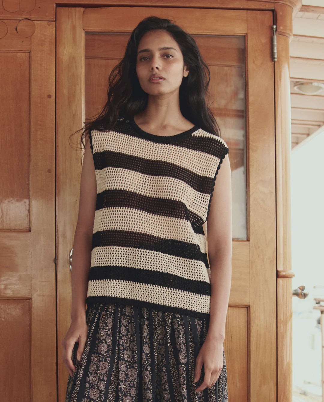 A woman with long dark hair stands in front of a wooden door, wearing The Crochet Sweater Shell by The Great Inc., paired with a patterned skirt. She looks directly at the camera with a calm expression.