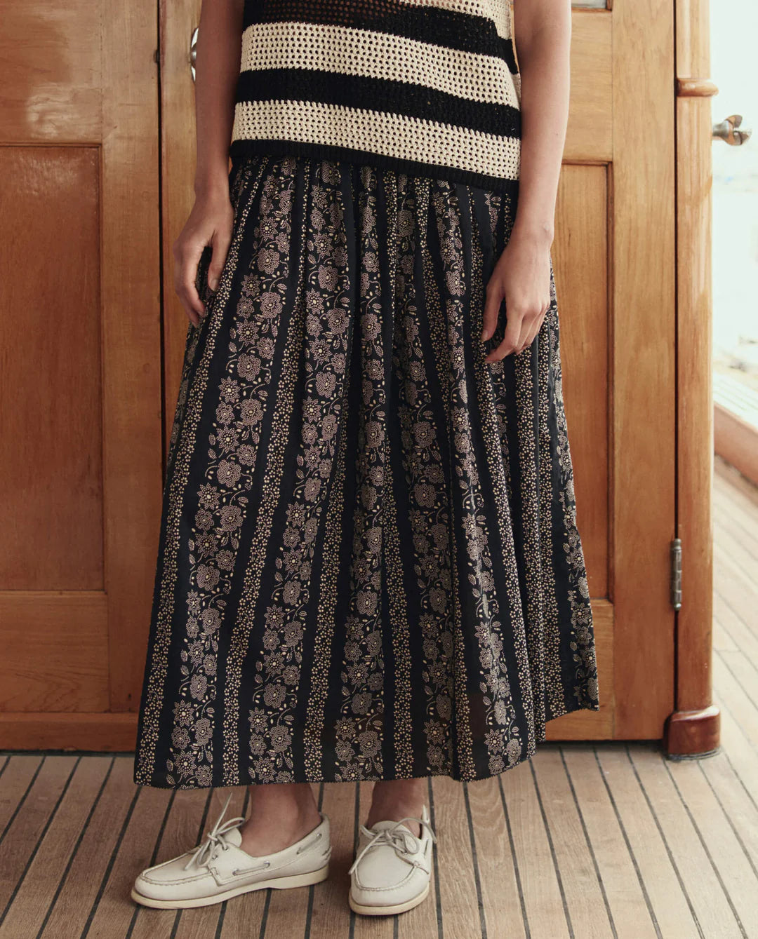 A person stands on a wooden floor wearing a black and cream striped sleeveless top, white boat shoes, and The Canary Skirt by The Great Inc., an airy pull-on skirt with floral and stripe patterns. The person's face is not visible.