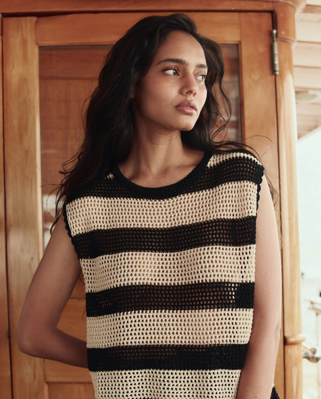 A woman with long dark hair stands indoors in front of a wooden door, wearing The Great Inc.'s Crochet Sweater Shell—a sleeveless black and beige striped crochet top with vintage sailor-inspired style. She gazes thoughtfully to the side.