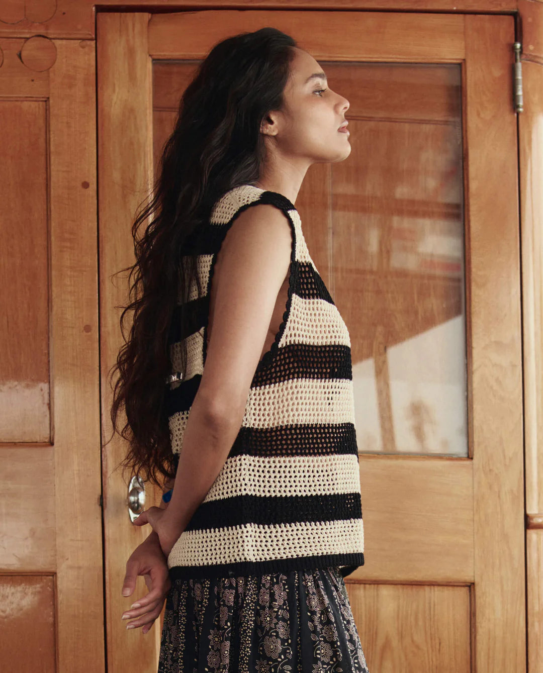 A woman with long dark hair stands in profile before wooden doors, wearing The Crochet Sweater Shell by The Great Inc., her hands clasped behind her back.