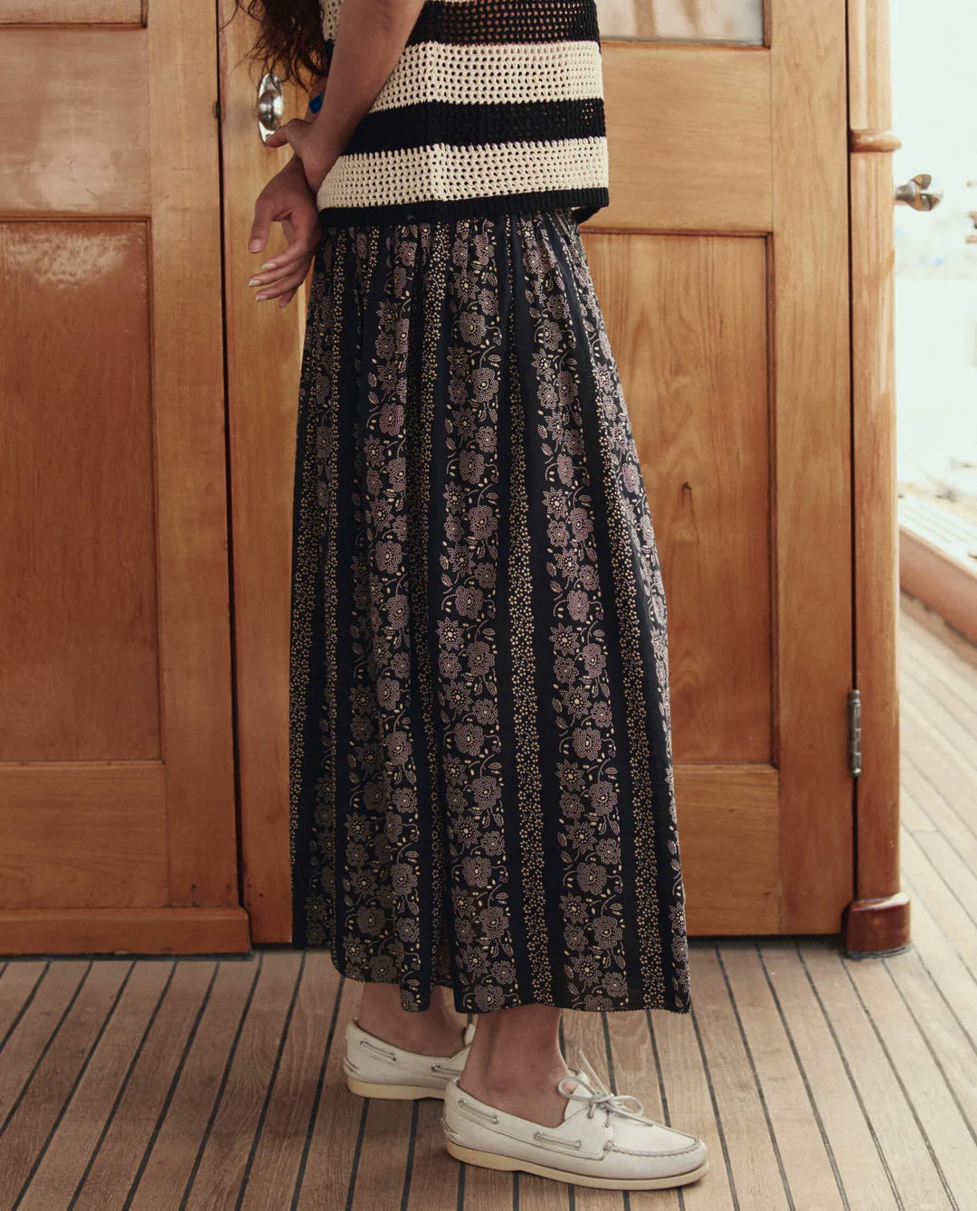 A person stands indoors on a wooden floor, wearing The Canary Skirt by The Great Inc.—an airy pull-on skirt with floral designs and vertical stripes—paired with a black and white striped sleeveless top and white boat shoes. Their face is out of the frame.