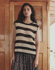 A woman with long dark hair stands in front of a wooden door, wearing The Crochet Sweater Shell by The Great Inc., paired with a patterned skirt. She looks directly at the camera with a calm expression.