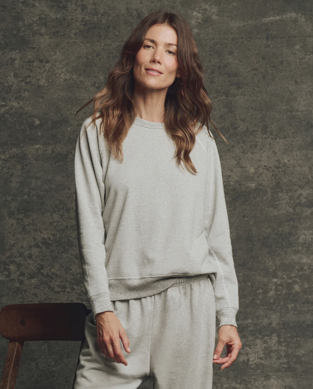 A woman with long brown hair wears The Fleece Rookie Sweatshirt by The Great Inc., paired with matching sweatpants, as she stands smiling slightly before a textured gray backdrop.