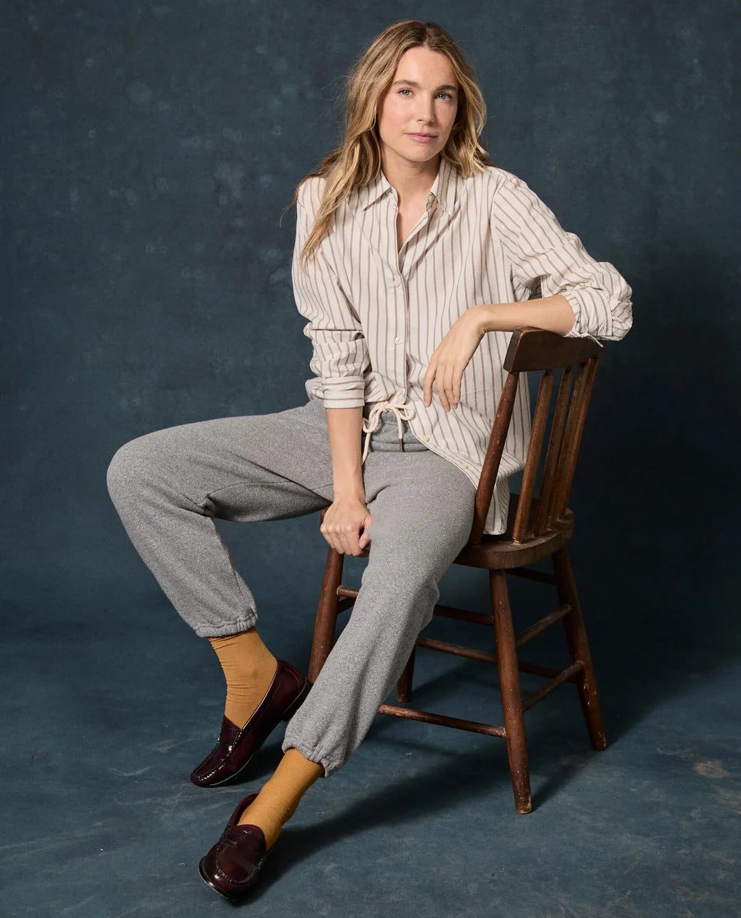 The Fleece Stadium Sweatpant on a seated woman wearing a striped shirt, mustard socks, and loafers in a wooden chair.