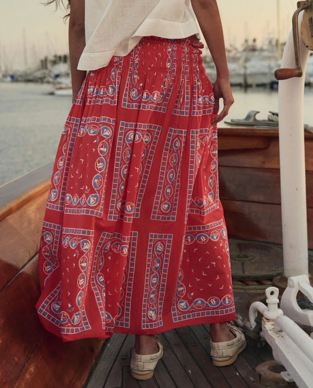 Wearing The Canary Skirt by The Great Inc.—a long, airy pull-on skirt with blue and white geometric and nautical patterns—a person stands on a wooden boat at sunset with a glowing marina in the background.