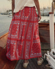 Wearing The Canary Skirt by The Great Inc.—a long, airy pull-on skirt with blue and white geometric and nautical patterns—a person stands on a wooden boat at sunset with a glowing marina in the background.