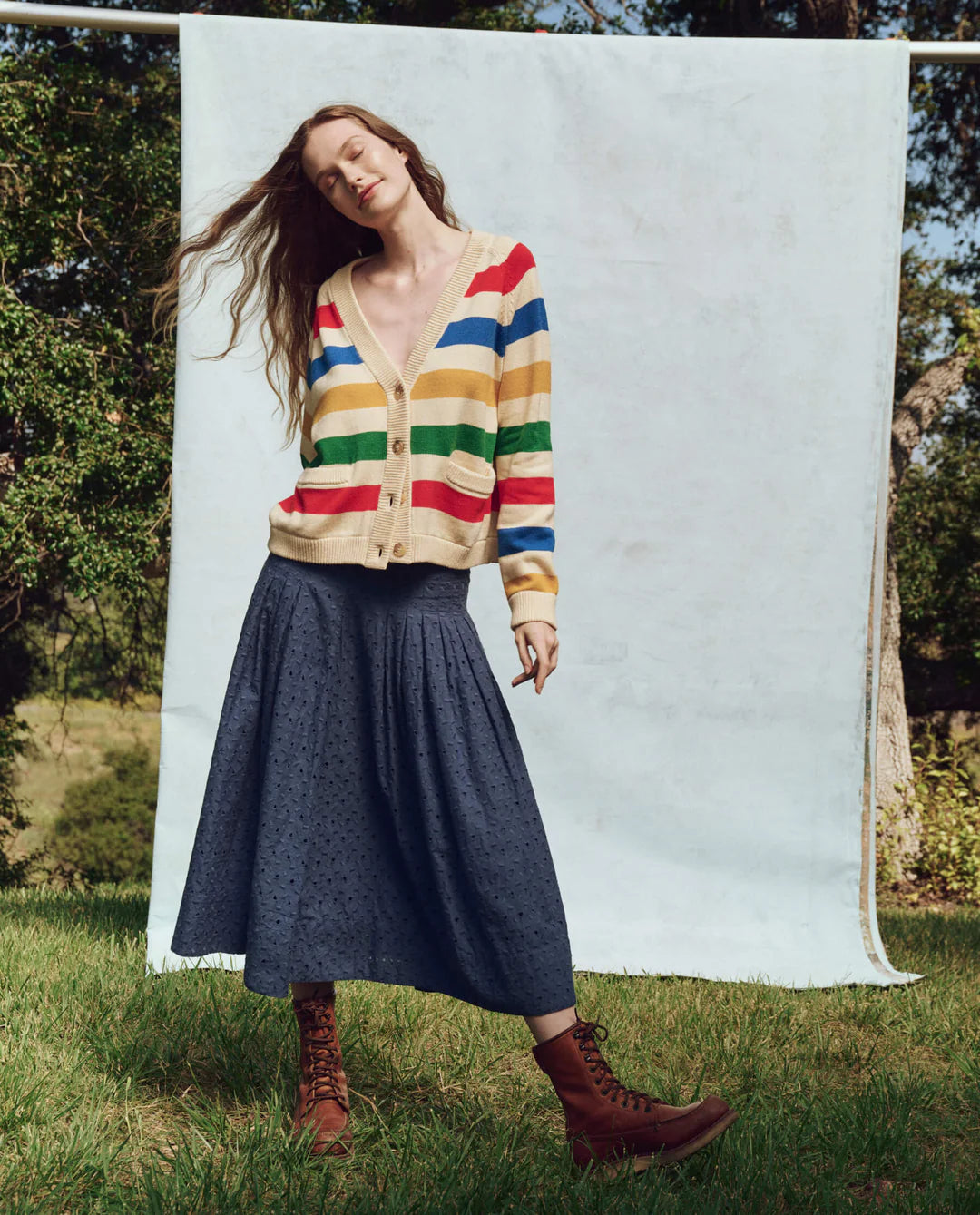 On sunny grass, a person wears a colorful striped cardigan, The Rhythm Skirt by The Great Inc. in long blue cotton eyelet style, and brown boots. They're framed by trees with a light blue backdrop, exuding Victorian charm.