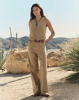 A woman stands outdoors among rocks and desert plants, wearing The Mill Pant by The Great Inc.—tan cotton herringbone workwear pants—with a matching vest, brown belt, and shoes. Mountains rise in the background.