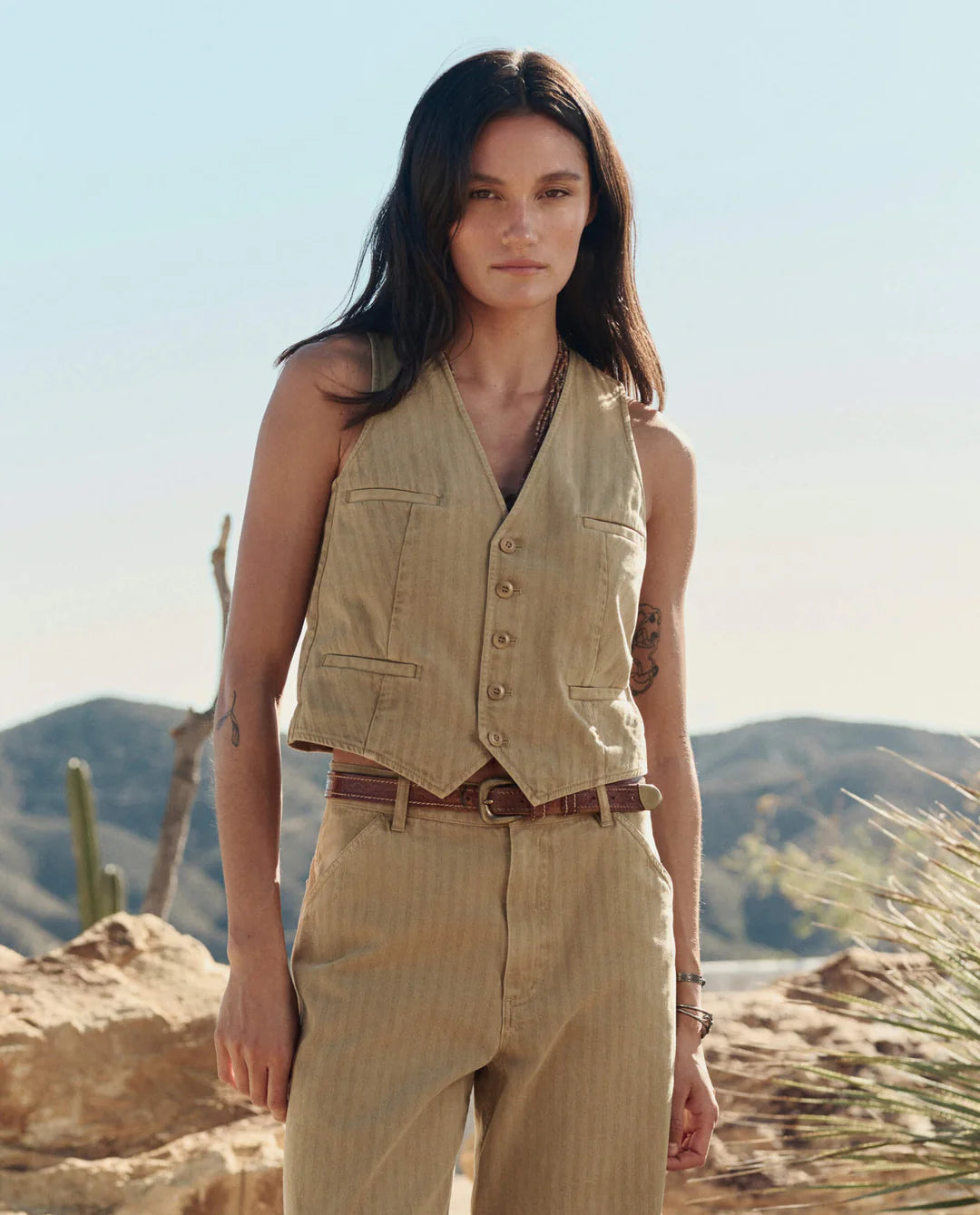 A person with long, dark hair stands in a desert landscape wearing The Suit Vest by The Great Inc., paired with matching pants and a brown belt. Cacti and rocky hills appear in the background beneath a clear blue sky.