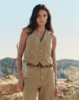 A person with long, dark hair stands in a desert landscape wearing The Suit Vest by The Great Inc., paired with matching pants and a brown belt. Cacti and rocky hills appear in the background beneath a clear blue sky.