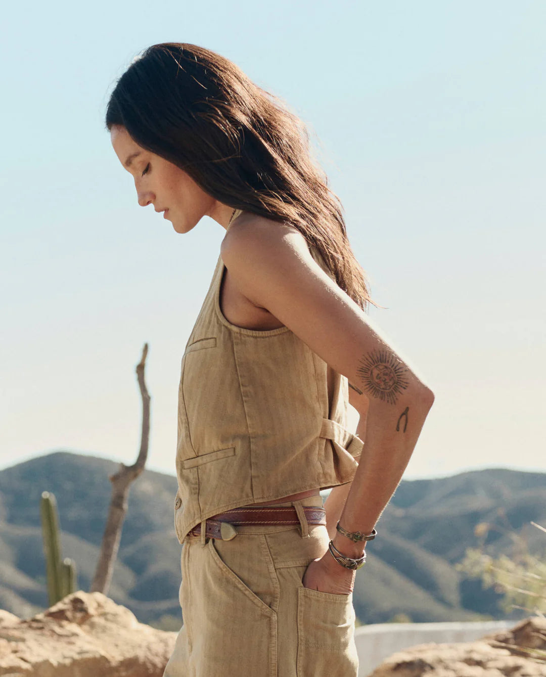 A woman with long brown hair and an upper arm tattoo stands outdoors in profile, wearing The Great Inc.'s The Suit Vest. Desert mountains and cacti are in the scenic background.