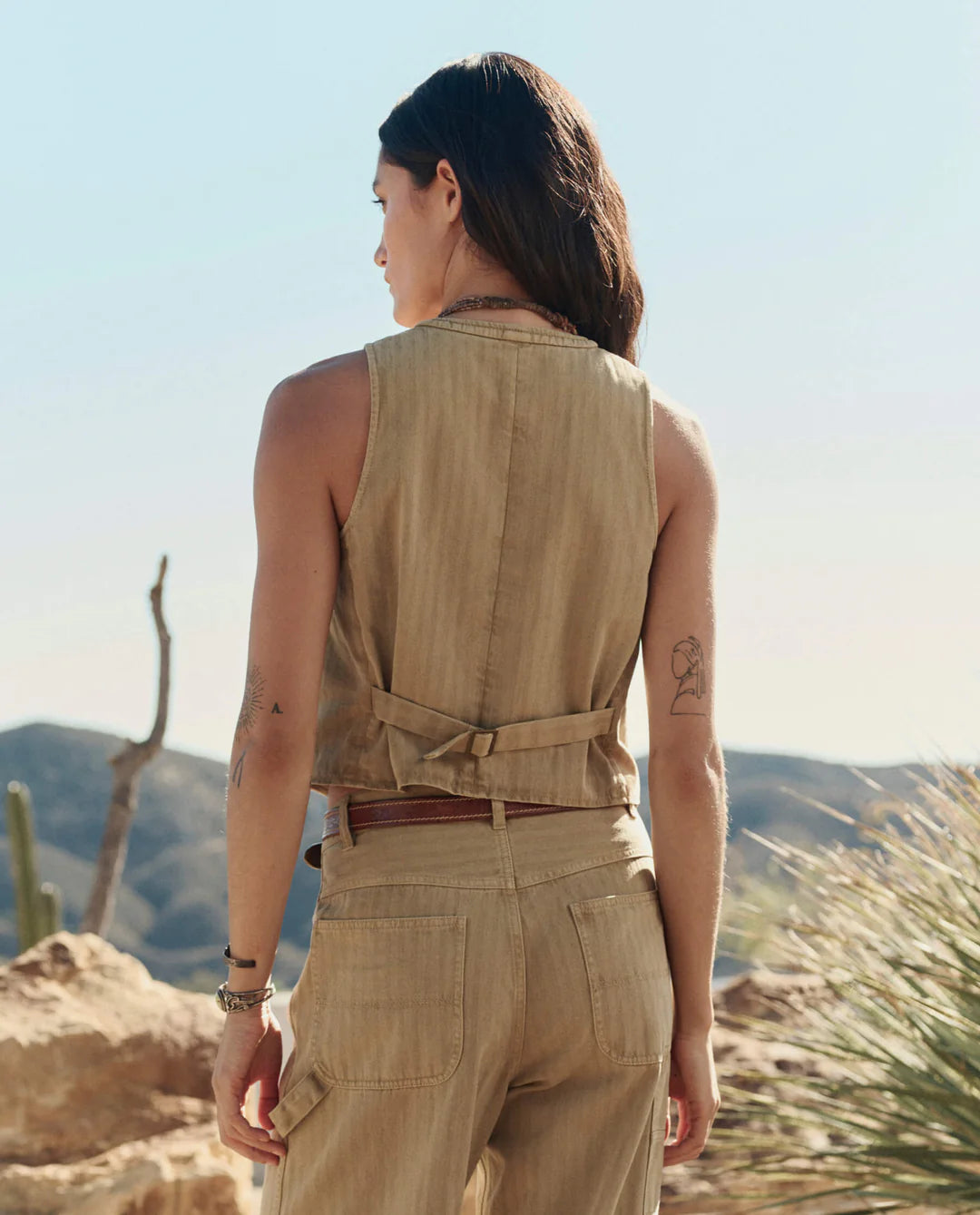 A woman stands outdoors with her back to the camera, wearing The Suit Vest and matching pants by The Great Inc. in light brown cotton herringbone, set against a desert landscape with cacti and rocky hills.