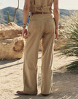 A person stands in a desert landscape, wearing The Mill Pant by The Great Inc.—loose, tan cotton herringbone workwear—paired with a matching sleeveless top. Their back is to the camera, with tattoos and bracelets visible. Desert plants and rocks fill the background.