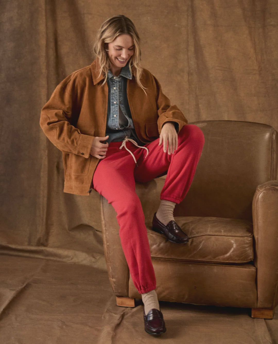 The Fleece Stadium Sweatpants in bright red worn with a brown suede jacket while seated in a leather armchair.