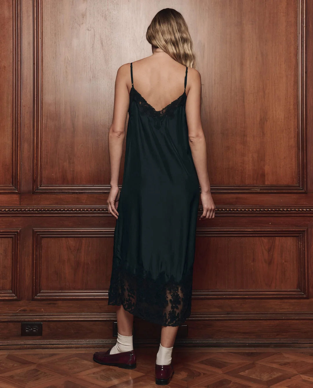 The Great Inc. black silk Legacy Dress with mesh lace, vintage-inspired, shown against a wood-paneled backdrop.