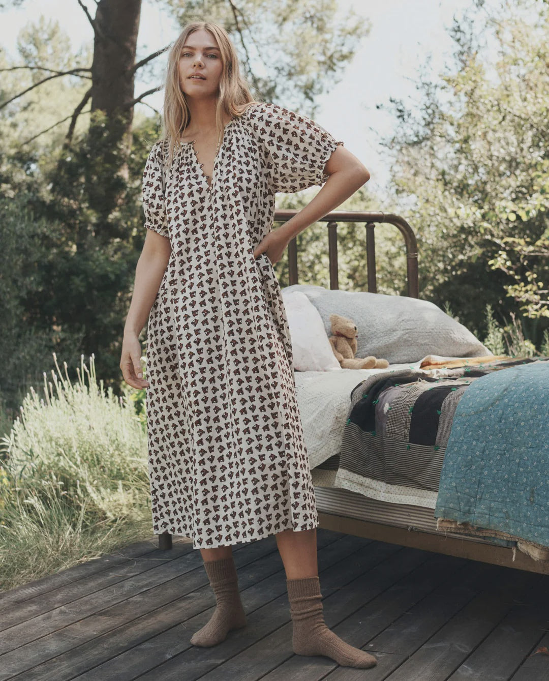 The Smocked Sleep Dress with brown bear print worn by a woman beside a metal bed frame in an outdoor setting.