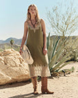 A woman wears The Great Inc.'s The Legacy Dress, a 1920s-style green slip with embroidered mesh lace, paired with brown cowboy boots. She stands in a desert setting holding a white hat amid rocks and agave under a clear sky.