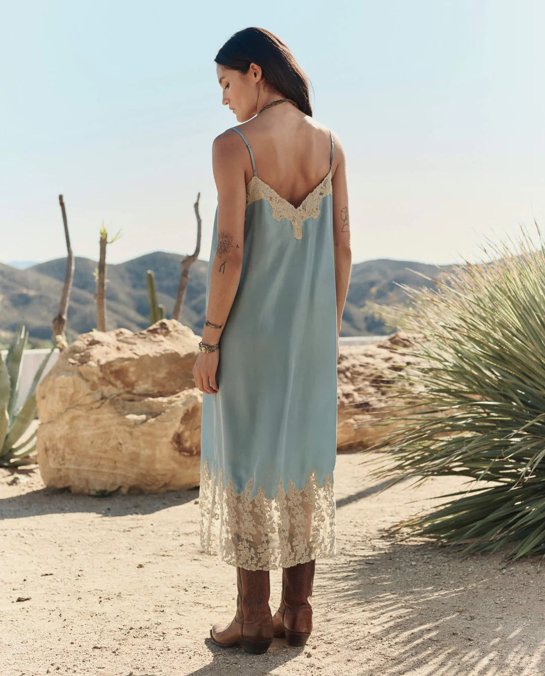 A woman stands outdoors in a desert landscape, facing away, wearing The Legacy Dress by The Great Inc.—a vintage-inspired piece with embroidered mesh lace—paired with brown boots. Cacti and dry mountains are visible under a clear sky.