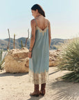 A woman stands outdoors in a desert landscape, facing away, wearing The Legacy Dress by The Great Inc.—a vintage-inspired piece with embroidered mesh lace—paired with brown boots. Cacti and dry mountains are visible under a clear sky.