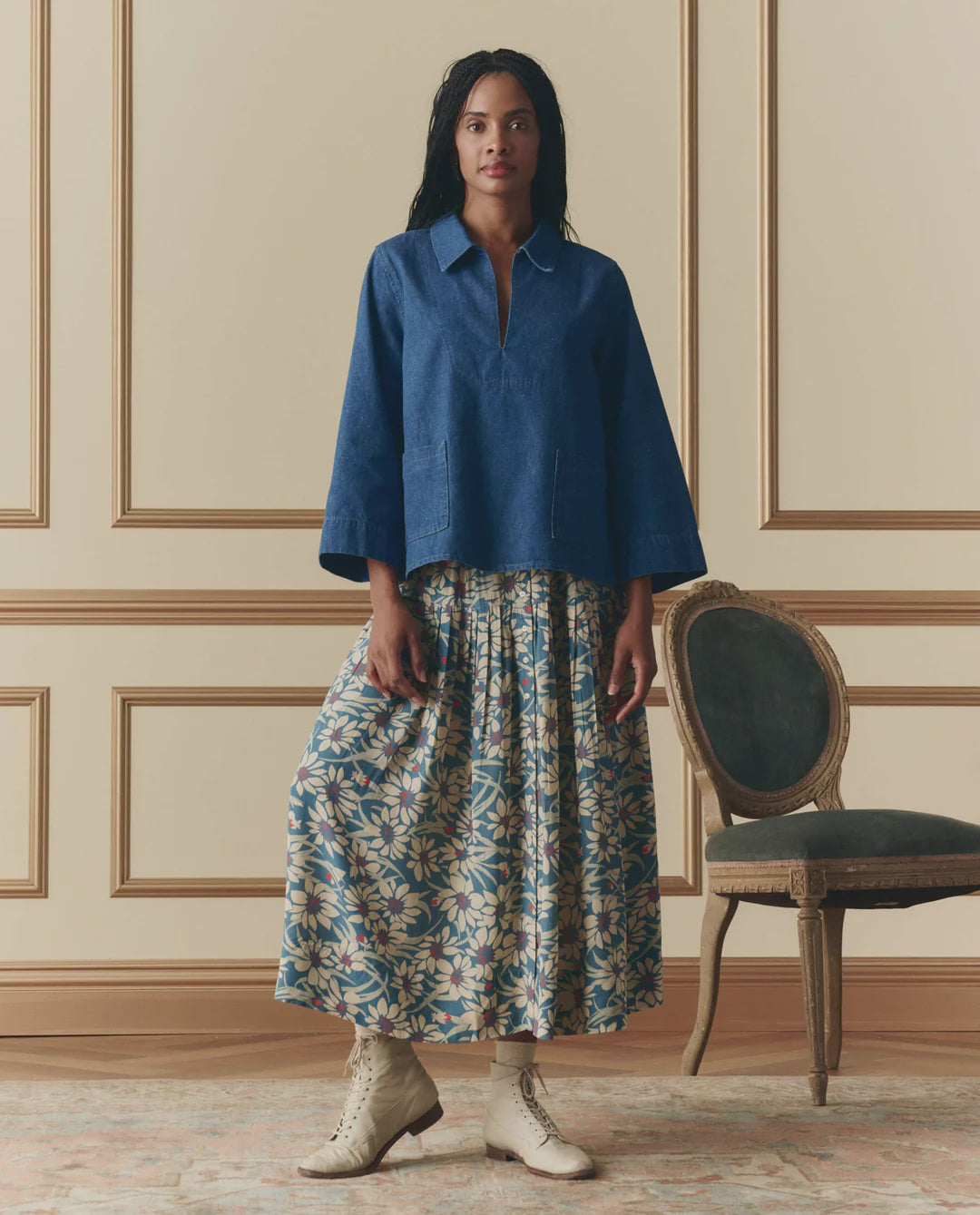 A woman stands indoors beside a vintage chair, wearing The Highland Skirt by The Great Inc.—a floral pleated skirt with an elastic waistband—paired with a loose blue top and light lace-up boots, against cream paneled walls and a patterned rug.