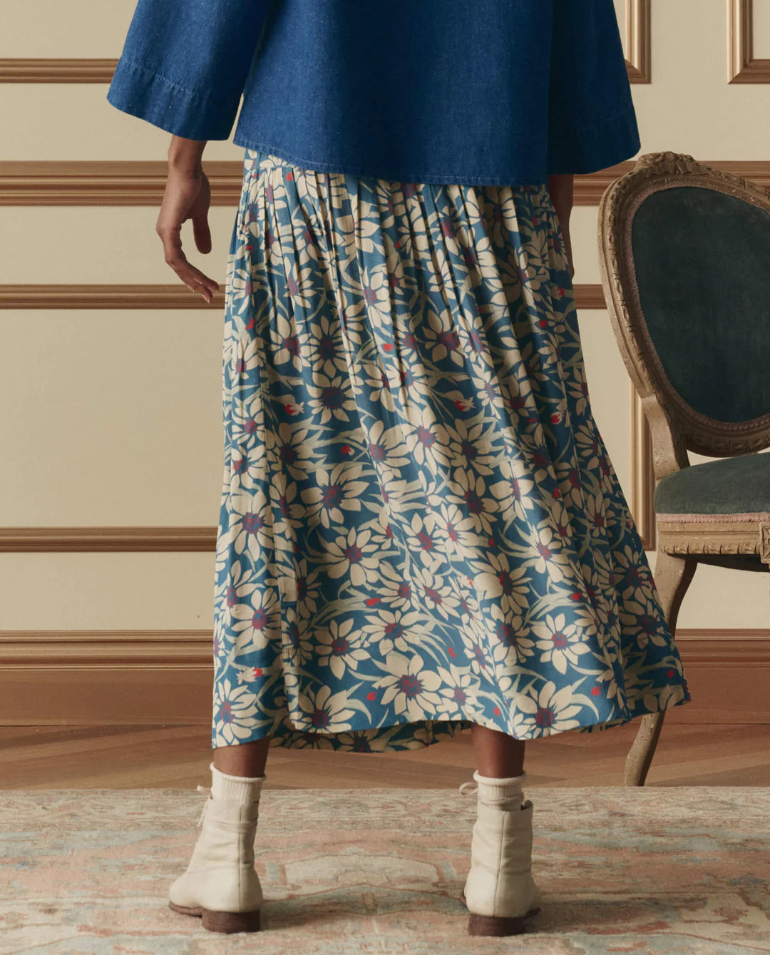 Indoors, a person wears a blue top with The Highland Skirt by The Great Inc.—a blue floral pleated piece featuring yellow and white flowers—paired with beige socks, white ankle boots, and a vintage-style chair nearby.