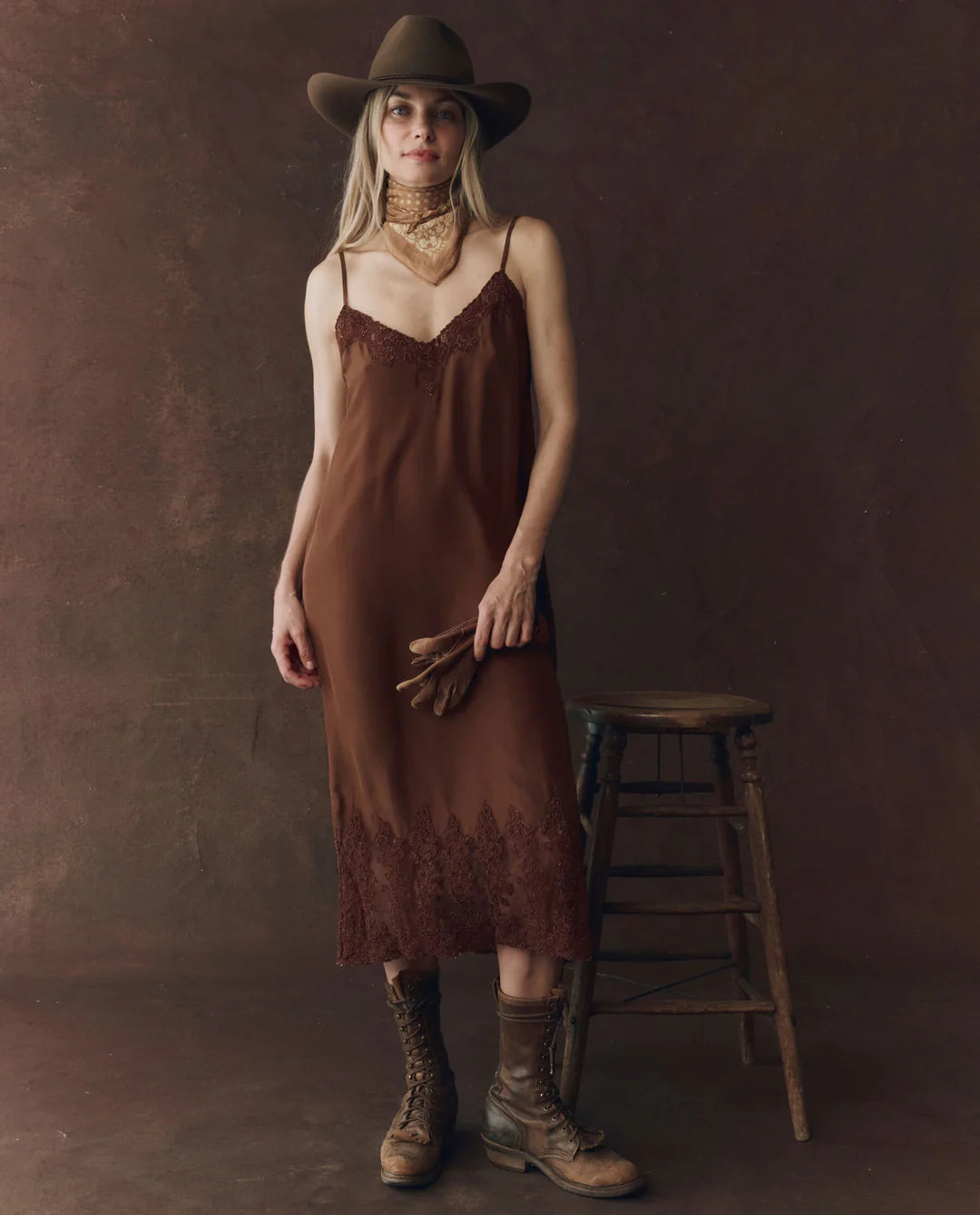 A woman models The Legacy Dress by The Great Inc.—a vintage-inspired brown silk dress—paired with a cowboy hat, boots, brown gloves, and a gold scarf beside a wooden stool, set against a dark brown backdrop.