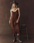 A woman models The Legacy Dress by The Great Inc.—a vintage-inspired brown silk dress—paired with a cowboy hat, boots, brown gloves, and a gold scarf beside a wooden stool, set against a dark brown backdrop.